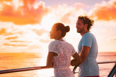 A full shot of a couple leaning on a boat railing against a bright sunset in the background